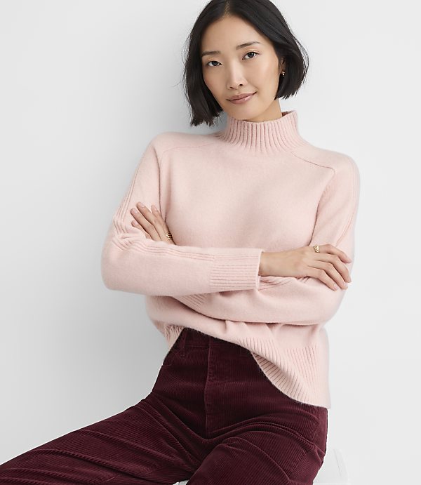 A woman wearing a light pink turtleneck sweater and maroon corduroy pants poses with crossed arms against a plain white background.