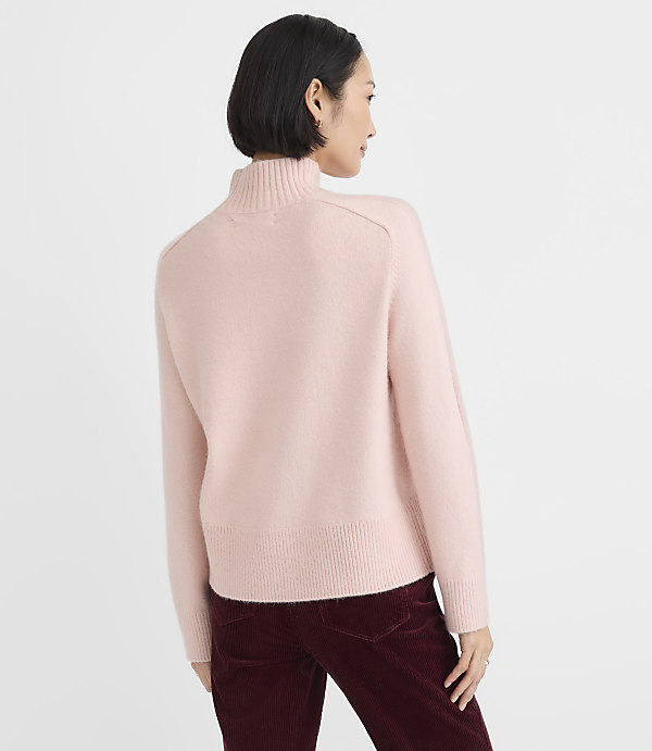 A woman wearing a light pink turtleneck sweater and dark burgundy corduroy pants stands against a plain white background.