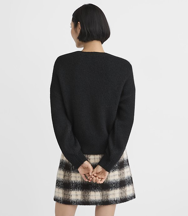 A woman wearing a black sweater and a plaid skirt stands with her back to the camera against a plain white background.