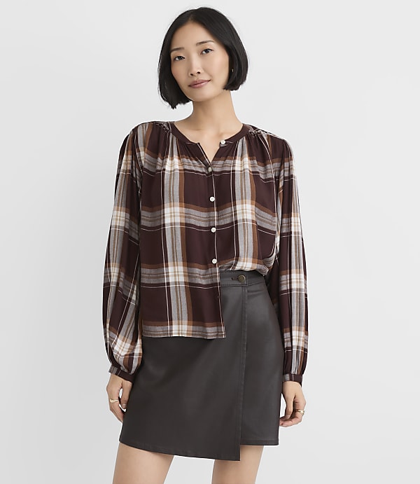 A woman wearing a brown plaid blouse and a dark grey faux-leather skirt stands against a plain white background.