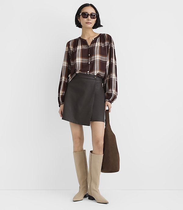 A woman wearing a plaid shirt, a dark grey skirt, and beige boots stands against a plain white background.