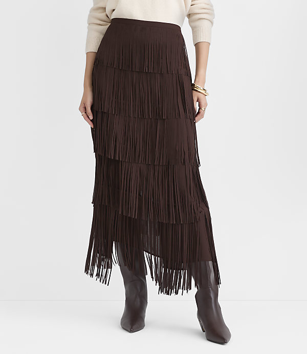 A person wearing a cream sweater, a tiered brown fringe skirt, and brown boots stands against a plain white background.
