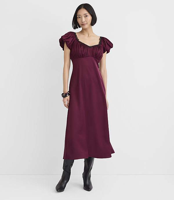 A woman wearing a maroon puff-sleeve dress and black boots stands against a plain white background.