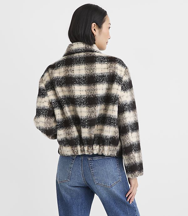 A woman wearing a brown and white checkered faux-fur jacket and blue jeans stands against a plain white background.