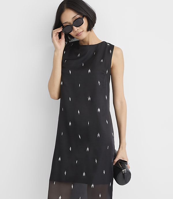 A woman wearing sunglasses and a black sleeveless dress with silver embellishments stands against a plain white background.