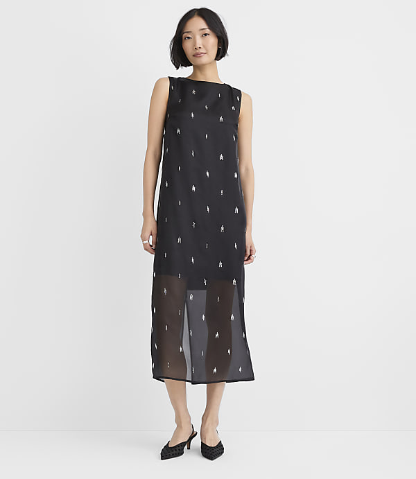 A woman wearing a black sleeveless midi dress with small white patterns and black heels stands against a plain white background.
