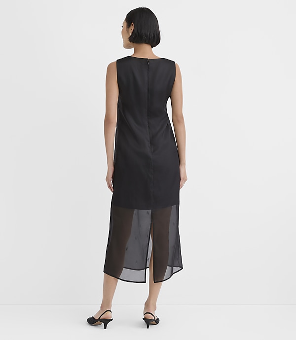 A woman wearing a sleeveless black dress with a sheer hem and black heels stands against a plain white background.