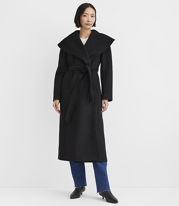 A woman wearing a long black belted coat and blue jeans stands against a plain white background.