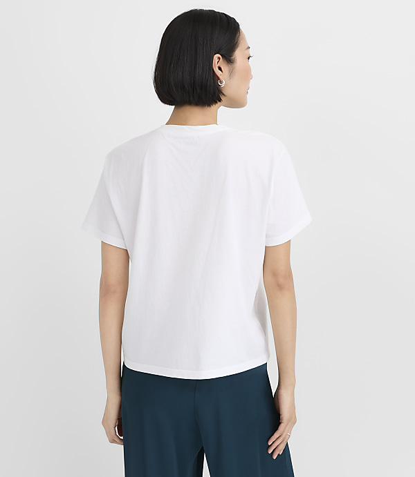 A woman with short black hair wearing a white t-shirt and dark teal pants stands against a plain white background.