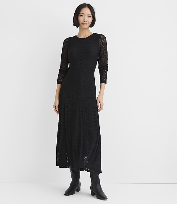 A woman wearing a long-sleeved black lace dress and black boots stands against a plain white background.