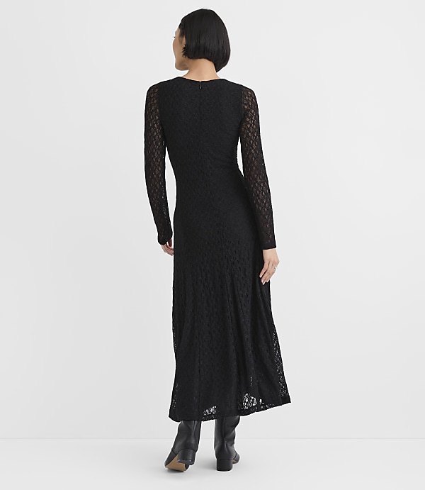 A woman wearing a long-sleeved black lace maxi dress and black boots stands with her back to the camera against a plain white background.