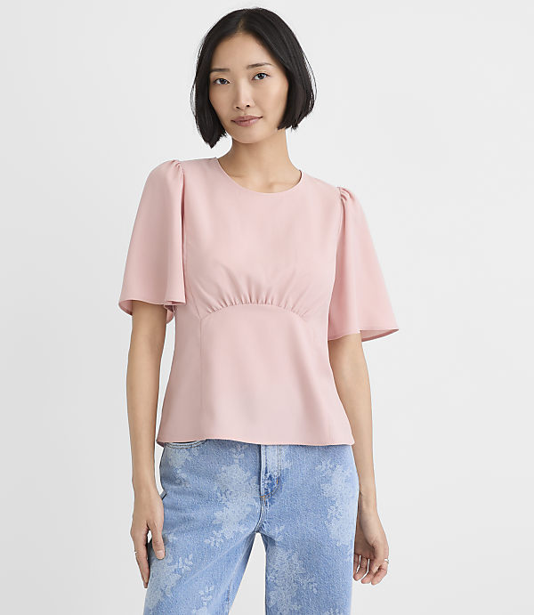 A woman wearing a light pink short-sleeved top and blue patterned jeans stands against a plain white background.