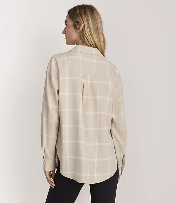 A woman wearing a beige plaid shirt and black pants stands against a plain light-colored background.