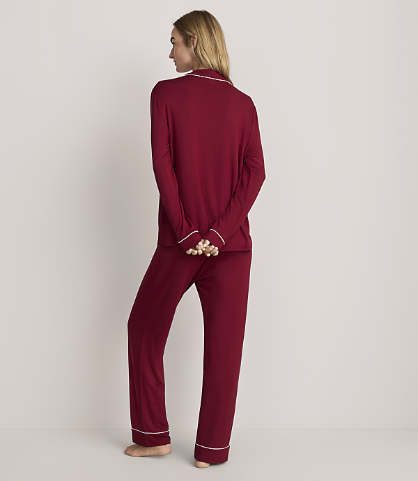 A woman stands with her back to the camera wearing burgundy pajamas with white trim against a plain, light-colored background.