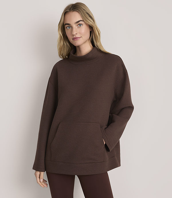 A blonde woman wearing a brown textured oversized tunic and leggings stands against a plain light-colored background.