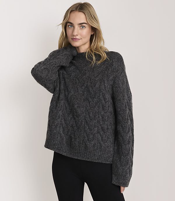 A blonde woman wearing a charcoal grey cable-knit sweater and black leggings stands against a plain white background.