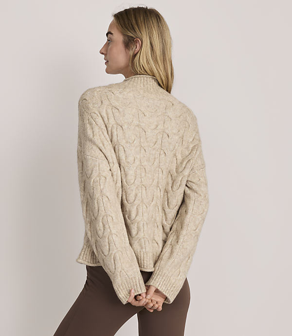 A woman wearing a beige cable-knit sweater and brown pants stands with her back to the camera against a plain light-colored background.