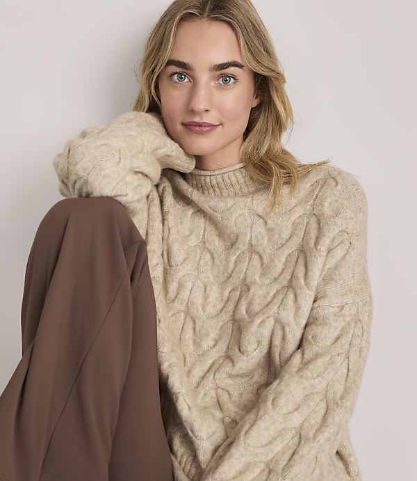 A woman wearing a beige cable-knit sweater and brown trousers poses against a plain, light-colored background.