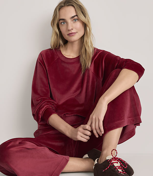 A woman wearing a red velvet lounge set and brown sneakers sits against a plain white background.