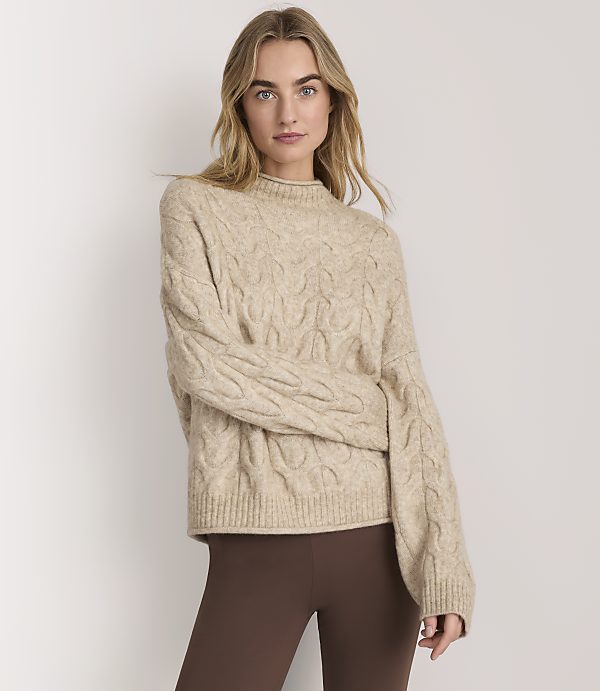 A woman wearing a beige cable-knit sweater and brown leggings stands against a plain white background.