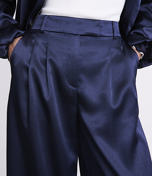A person wears navy blue satin pleated trousers and a dark jacket over a white top against a plain light background.
