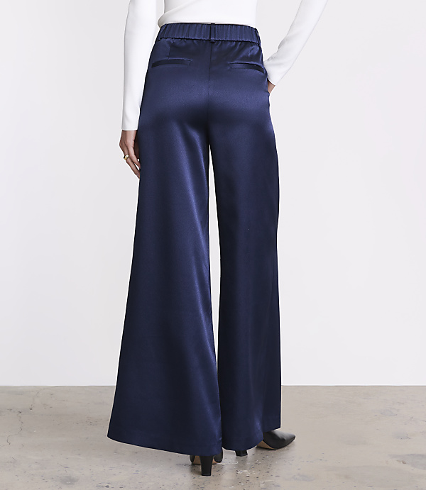 A person wearing a white long-sleeved top and wide-leg navy blue satin trousers stands against a plain white background.