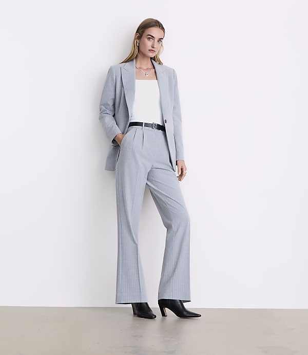 A woman wearing a light blue pinstripe suit and black heels stands against a plain white wall.