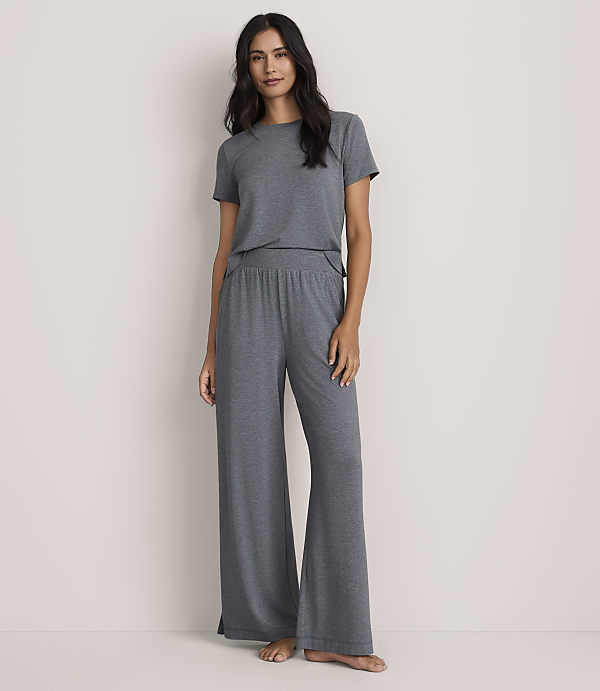 A woman wearing a grey short-sleeved t-shirt and matching wide-leg trousers stands against a plain, light-colored wall.