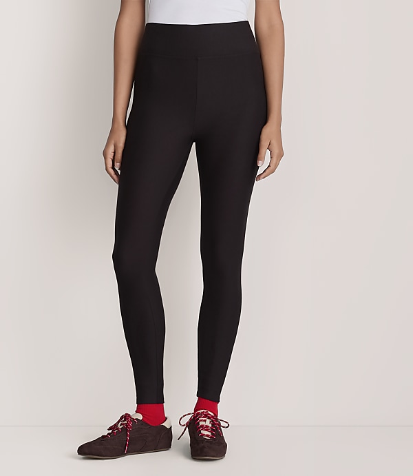 A person wearing a white top, black leggings, red socks, and brown sneakers stands against a plain white background.