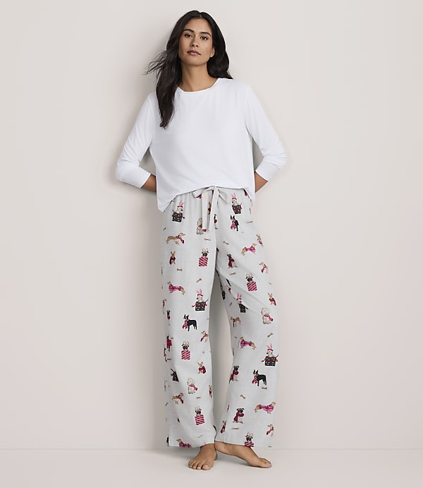 A woman wearing a white long-sleeved shirt and patterned pajama pants stands against a plain, light-colored background.