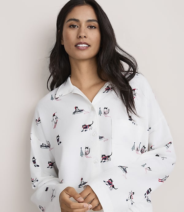 A woman with long dark hair wears a white button-down shirt featuring cat illustrations against a plain, light-colored background.