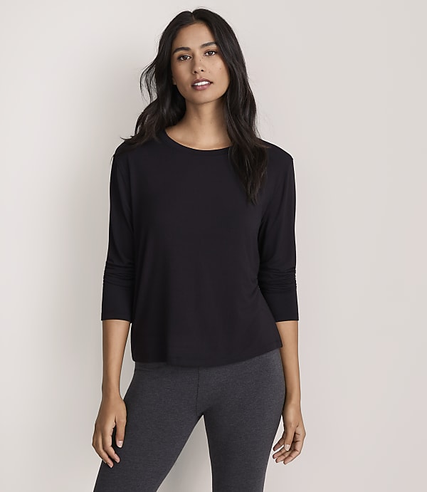 A woman wearing a black long-sleeved top and dark grey leggings stands against a plain, light-colored background.