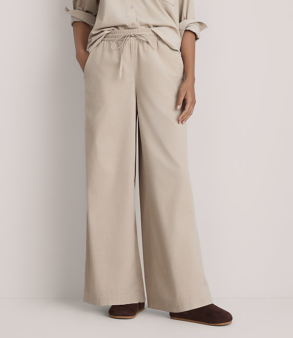 A person wearing a beige button-down shirt and wide-leg beige trousers stands against a plain white background.