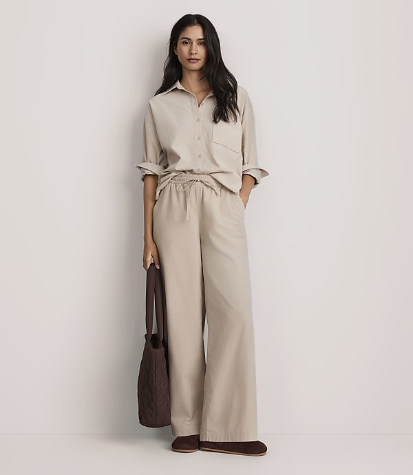 A woman wearing a beige button-down shirt and matching wide-leg trousers stands against a plain white background while holding a brown handbag.