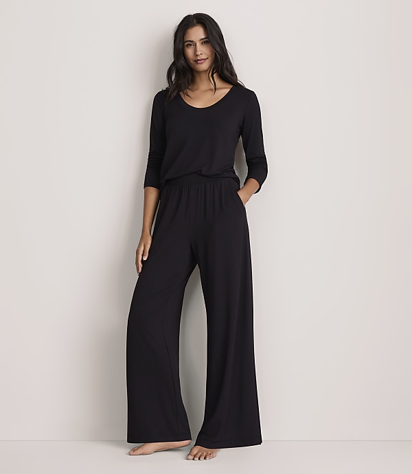 A woman wearing a black long-sleeved top and wide-leg black trousers stands against a plain, light-colored wall.
