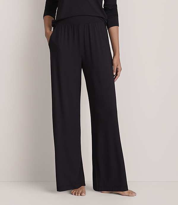 A person wearing a black long-sleeved top and wide-leg black trousers stands against a plain, light-colored wall.