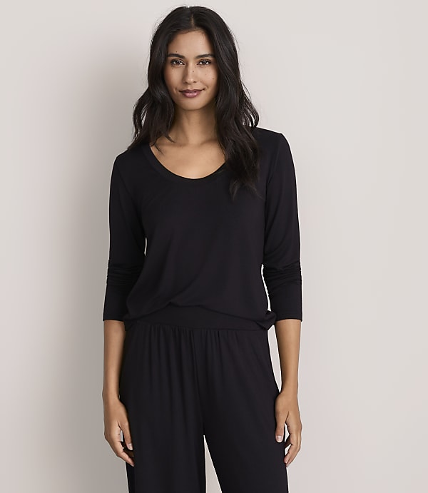 A woman with long dark hair wearing a black long-sleeved top and matching pants stands against a plain, light-colored background.