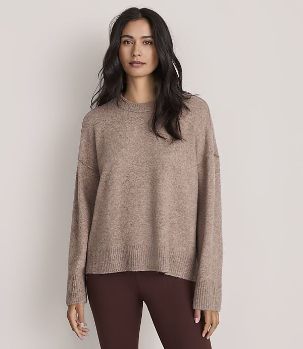A woman wearing a brown oversized sweater and dark brown leggings stands against a plain, light-colored background.
