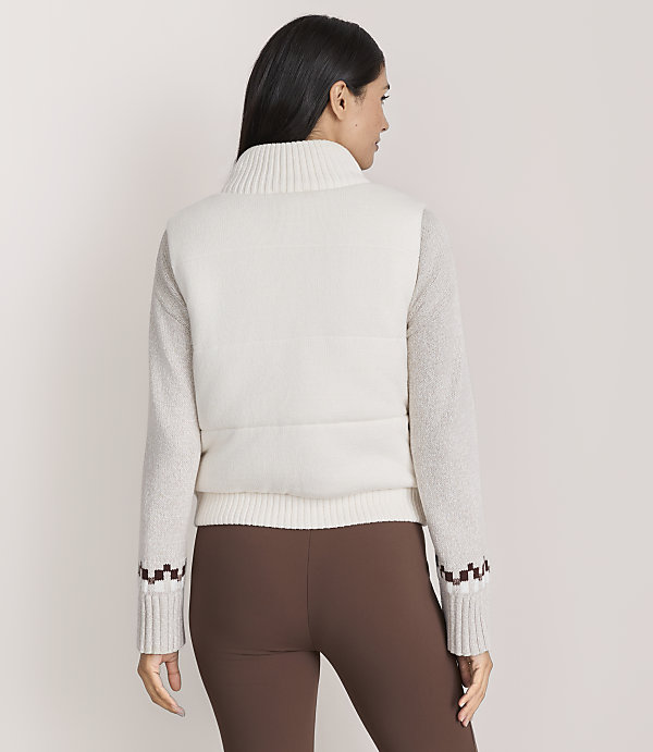 A woman wearing a white textured sweater and brown leggings stands with her back to the camera against a plain light-colored background.