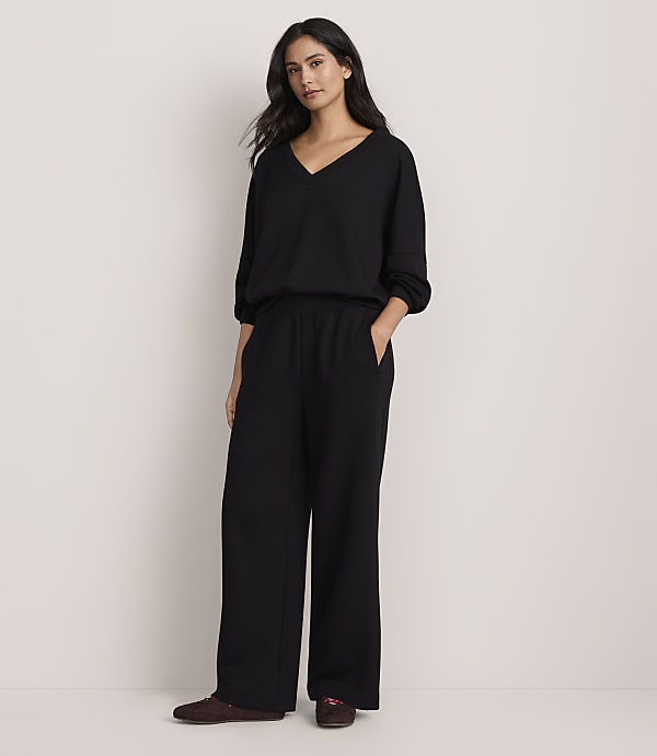 A woman wearing a black V-neck top and wide-leg black trousers stands against a plain, light-colored background.