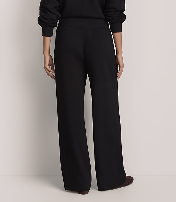 A person stands with their back to the camera wearing black wide-leg trousers and a black top against a plain white background.