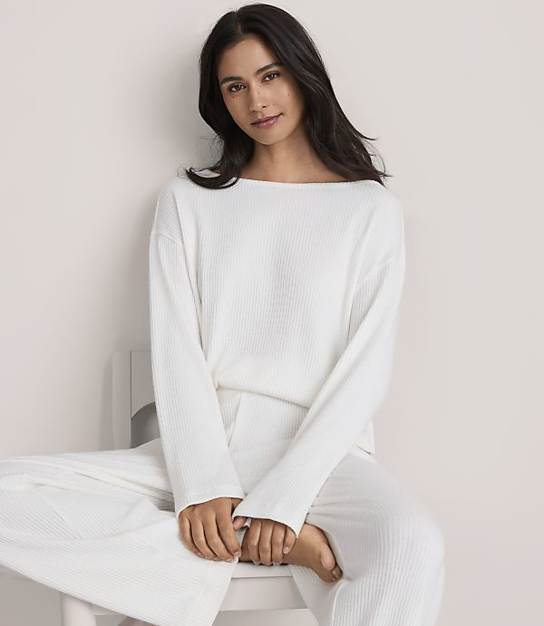 A woman wearing a white ribbed lounge set sits on a white chair against a plain, light-colored wall.