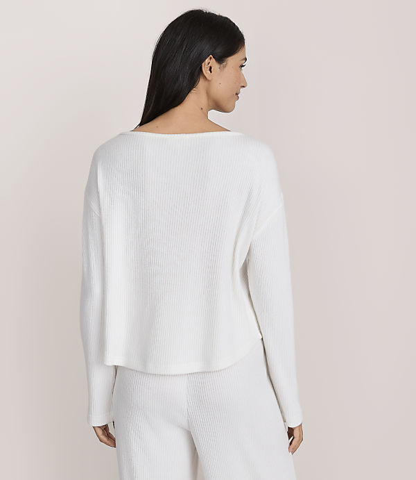A woman with long dark hair wearing a white ribbed long-sleeved top and matching pants stands against a plain light-colored background.