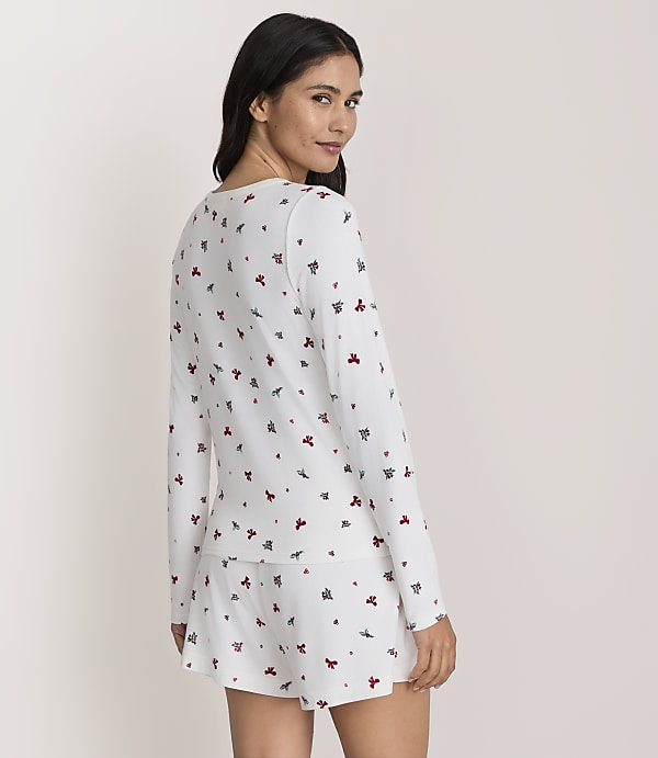 A woman wearing white pajamas with a small red and green pattern stands against a plain, light-colored background.