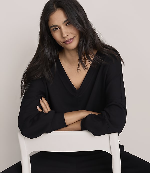 A woman with long dark hair wearing a black V-neck top leans on a white chair against a plain light-colored background.