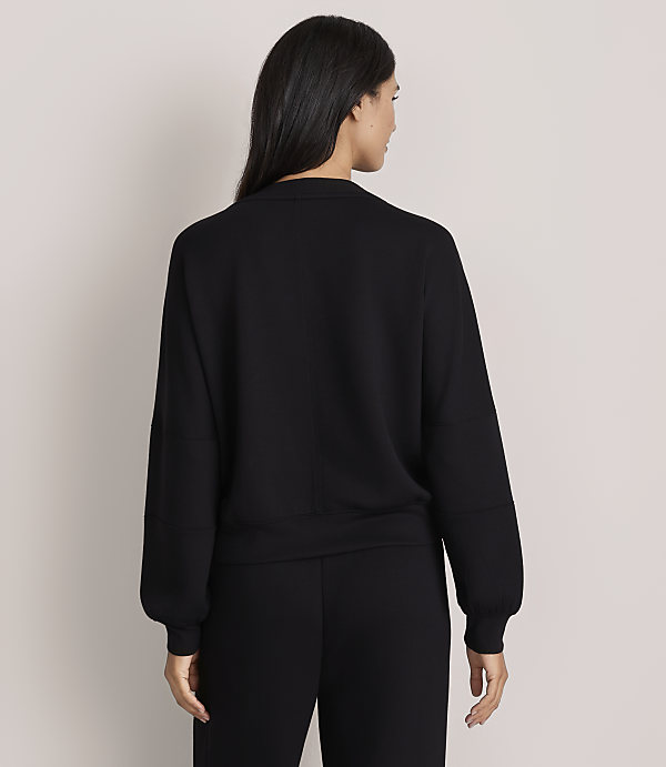 A woman with long dark hair stands with her back to the camera wearing a black long-sleeved top and black pants against a plain light-colored background.