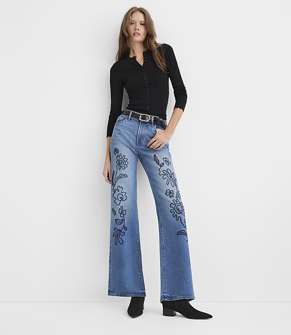 A woman wearing a black long-sleeved top and blue flared jeans with floral embroidery stands against a plain white background.
