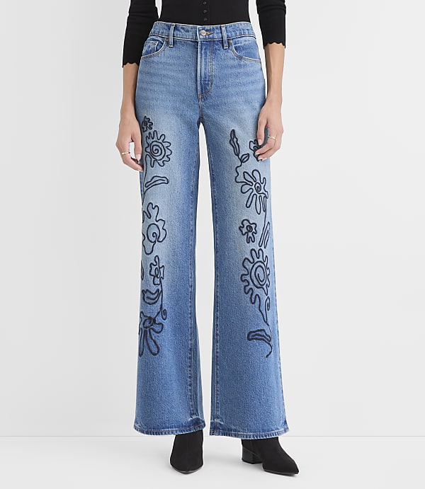 A person wears blue wide-leg jeans with black floral embroidery against a plain white background.