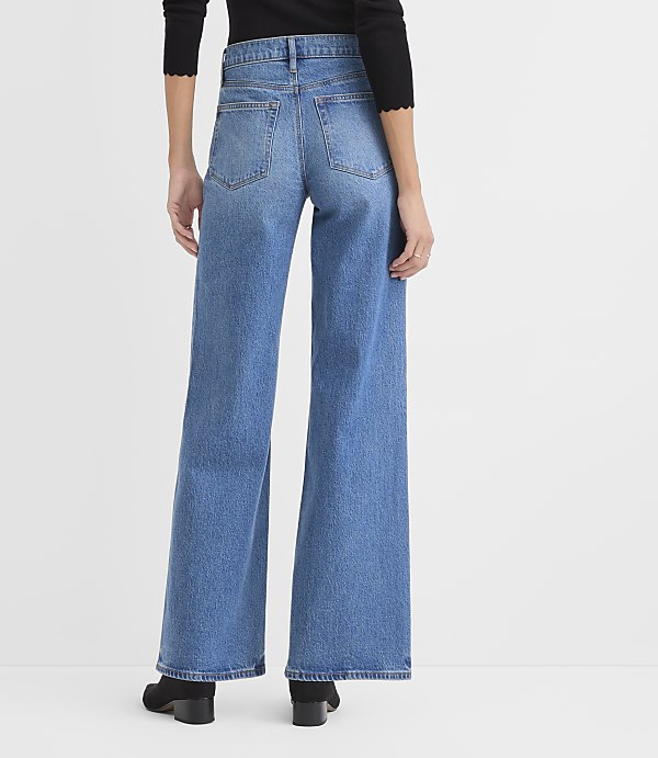 A person wearing a black top and wide-leg blue jeans stands against a plain white background.