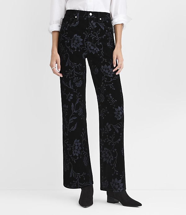 A person wears black floral velvet trousers, a white shirt, and black boots against a plain white background.